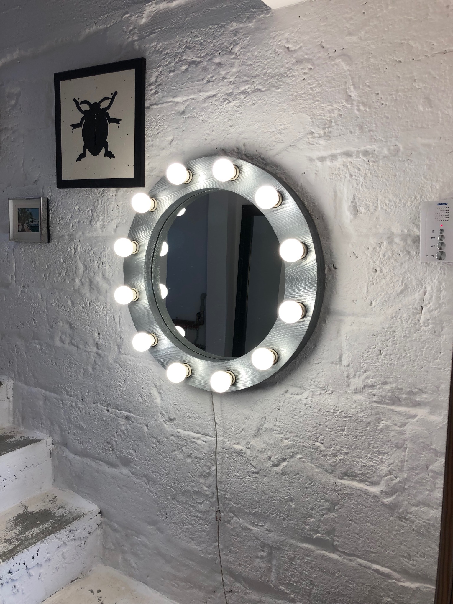 Okrągłe lustro z oświetleniem LED, zamontowane na białej, teksturowanej ścianie z cegły. Obok wisi czarno-biała grafika owada i domofon.