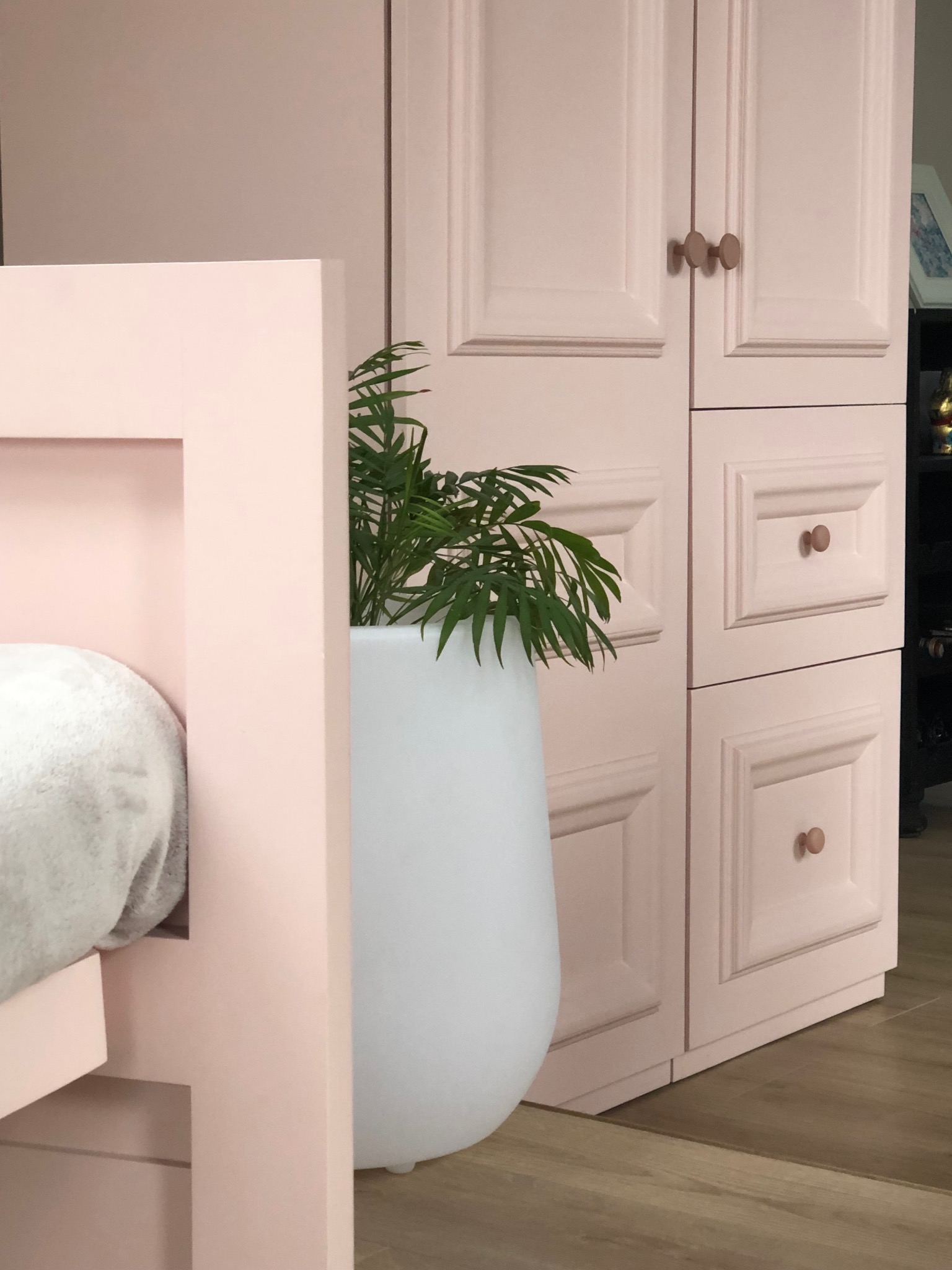 Różowa szafa z frezowanymi frontami i okrągłymi uchwytami, obok łóżko i roślina w białej donicy. Drewniana podłoga.