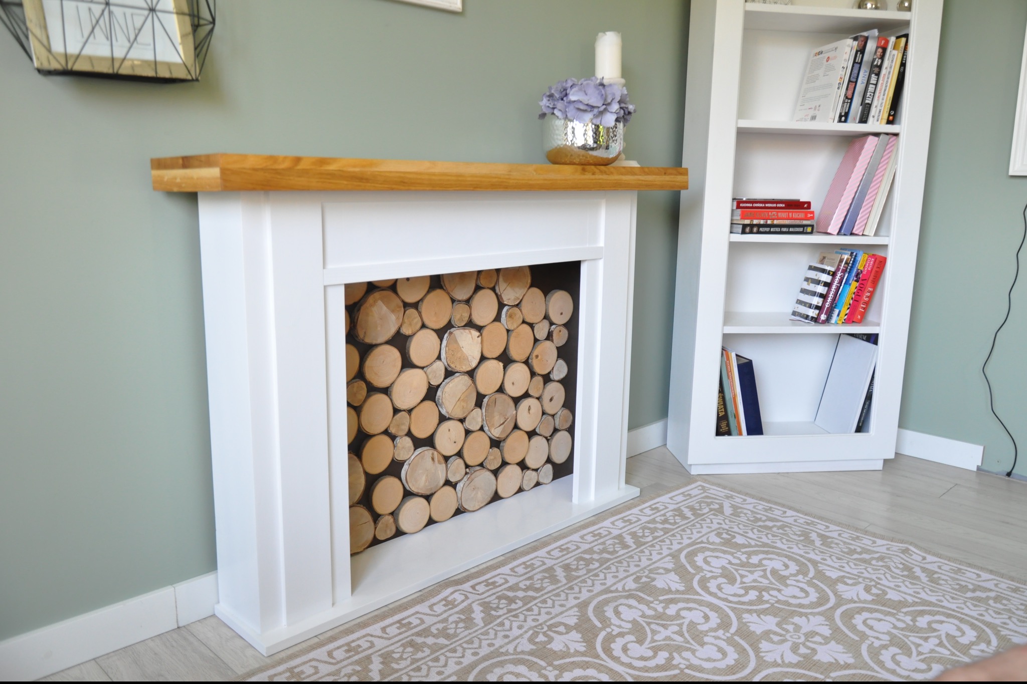 Biały, dekoracyjny kominek z drewnianym blatem, wypełniony pociętymi kawałkami drewna. W tle regał z książkami i dywan w kwiatowy wzór.