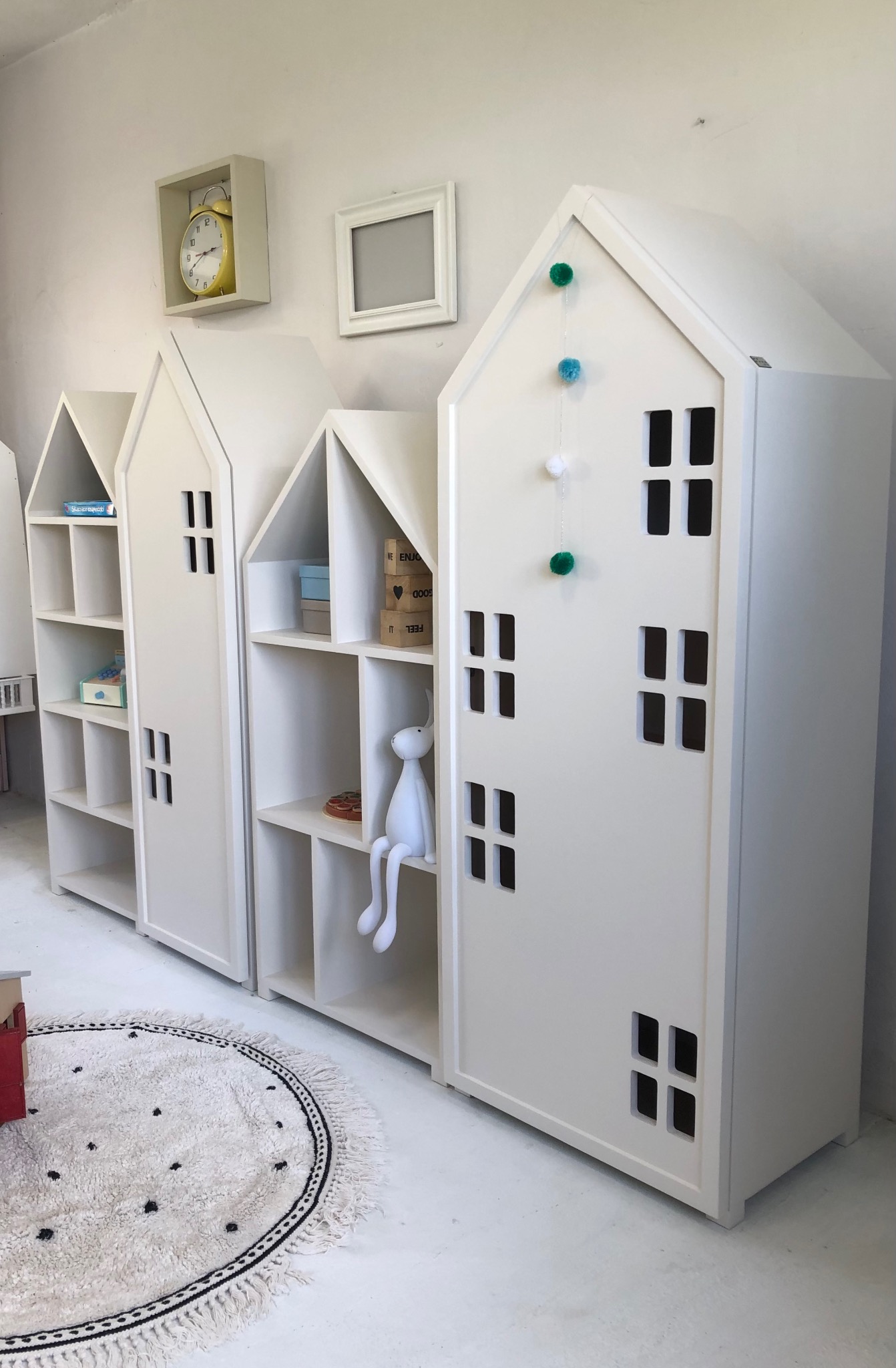 Białe regały w kształcie domków dziecięcych z dekoracjami, dywanik w kropki. Nietypowe meble do pokoju dziecięcego, nawiązujące do architektury.