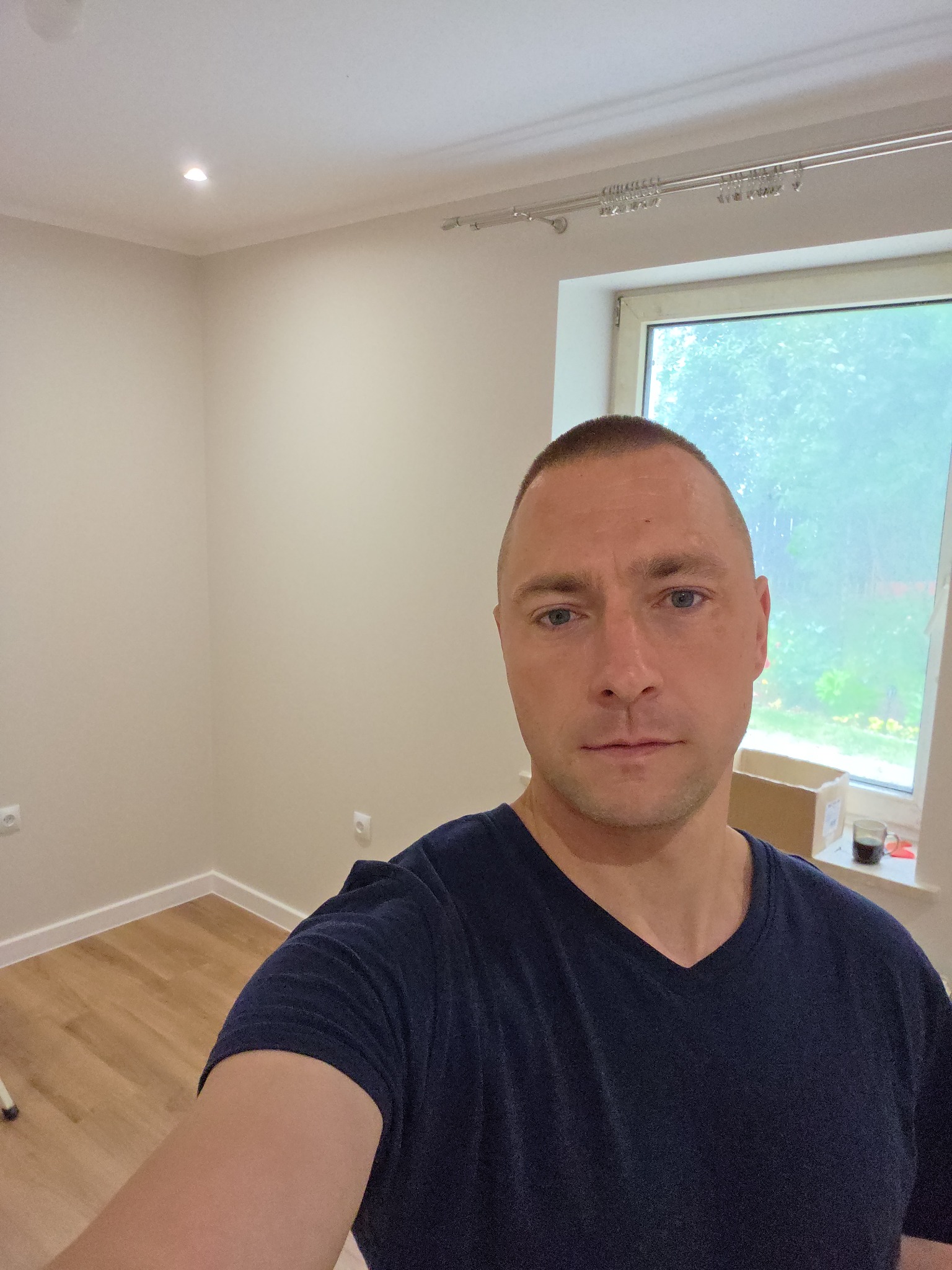 Jasne, puste pomieszczenie z oknem i parkietem. Mężczyzna w niebieskiej koszulce robi selfie na tle świeżo odmalowanych ścian.