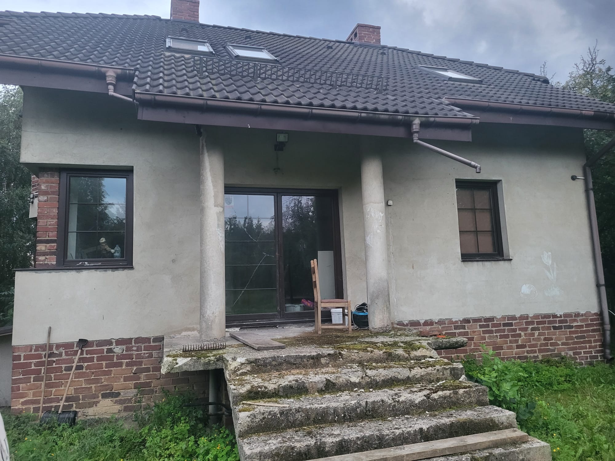 Dom z szarą elewacją, widoczne naprawy murarskie na fundamencie z cegły, schody wejściowe z betonu pokryte mchem. Okna brązowe, dachówka szara.