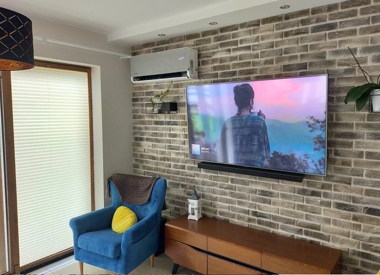 Szary klimatyzator ścienny w salonie z ceglaną ścianą, telewizorem i niebieskim fotelem. Nowoczesny design. Roślina doniczkowa po prawej stronie.