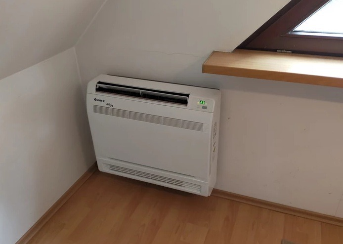 Biała klimatyzacja podłogowa Gree, zamontowana w rogu pomieszczenia ze skośnym sufitem i drewnianą podłogą. Widoczny wyświetlacz z temperaturą 23 stopnie.