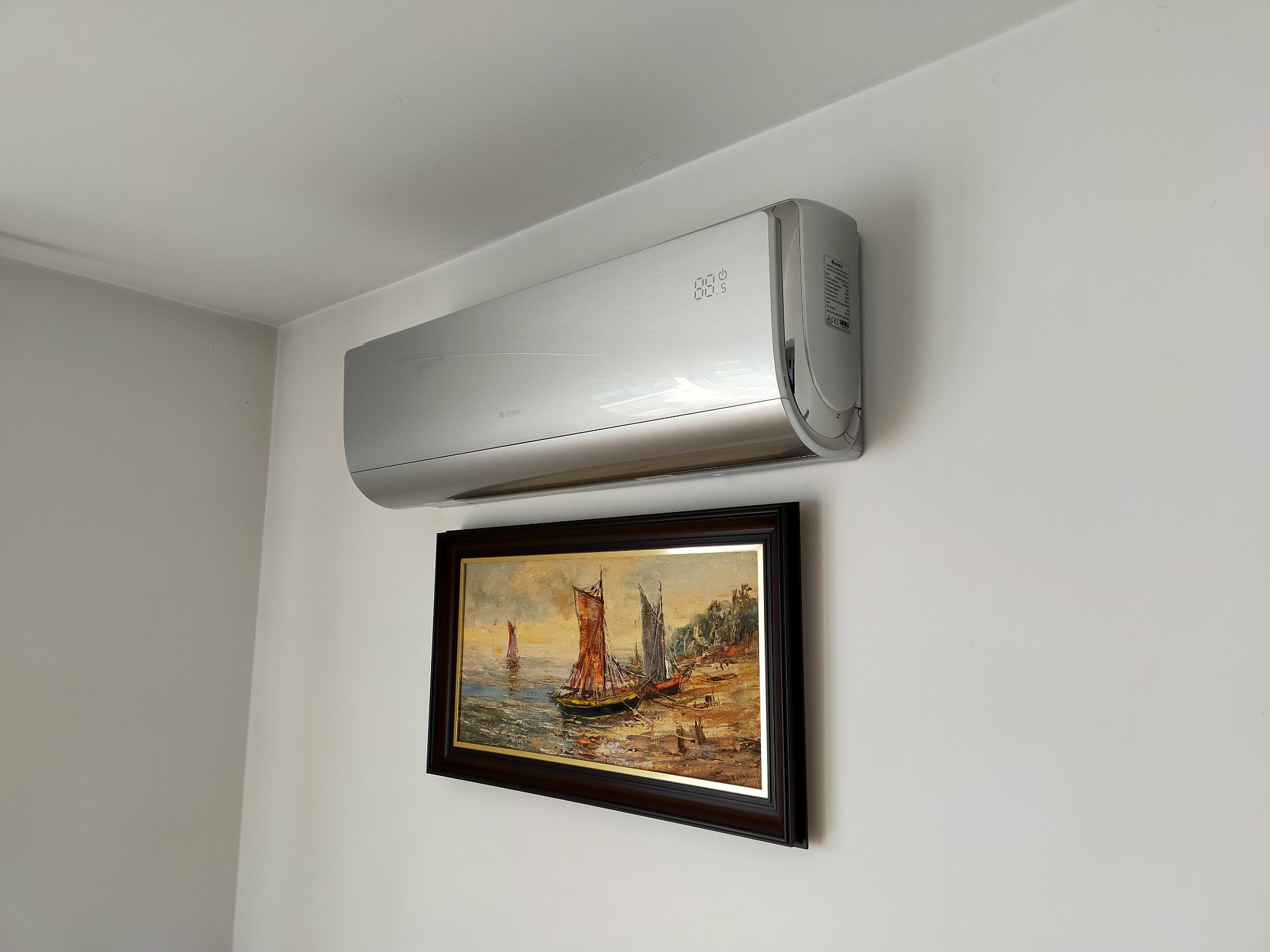 Srebrny klimatyzator ścienny marki Gree, zamontowany nad obrazem z motywem żaglówek. Biała ściana, widoczny narożnik pokoju. Nowoczesny design.
