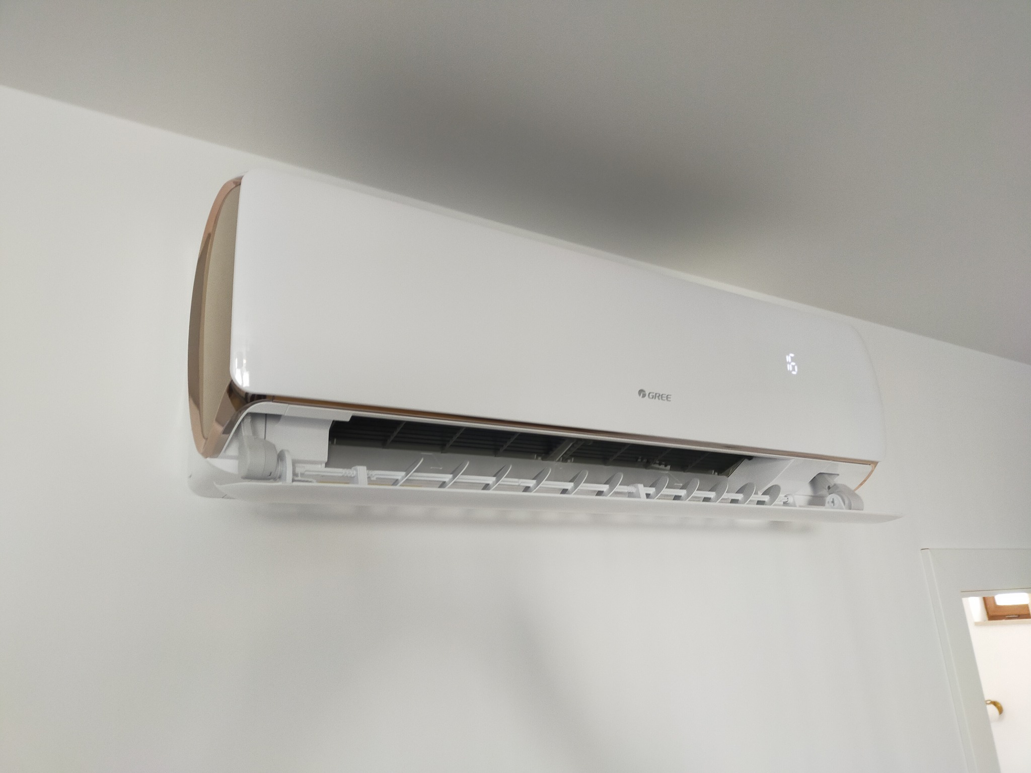 Nowoczesny, biały klimatyzator GREE z elementami w kolorze miedzianym, zamontowany na białej ścianie w jasnym pomieszczeniu. Widoczne otwarte klapy wentylacyjne.