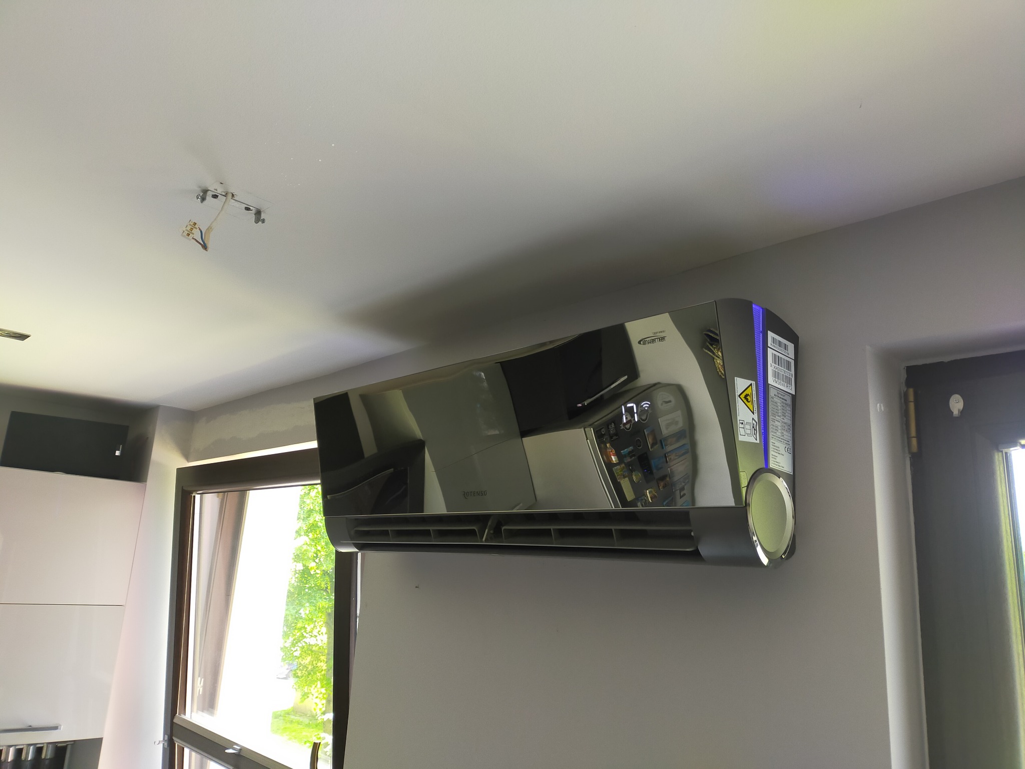 Błyszczący klimatyzator Rotenso zamontowany na szarej ścianie w pomieszczeniu z oknem i widocznym fragmentem instalacji elektrycznej na suficie.