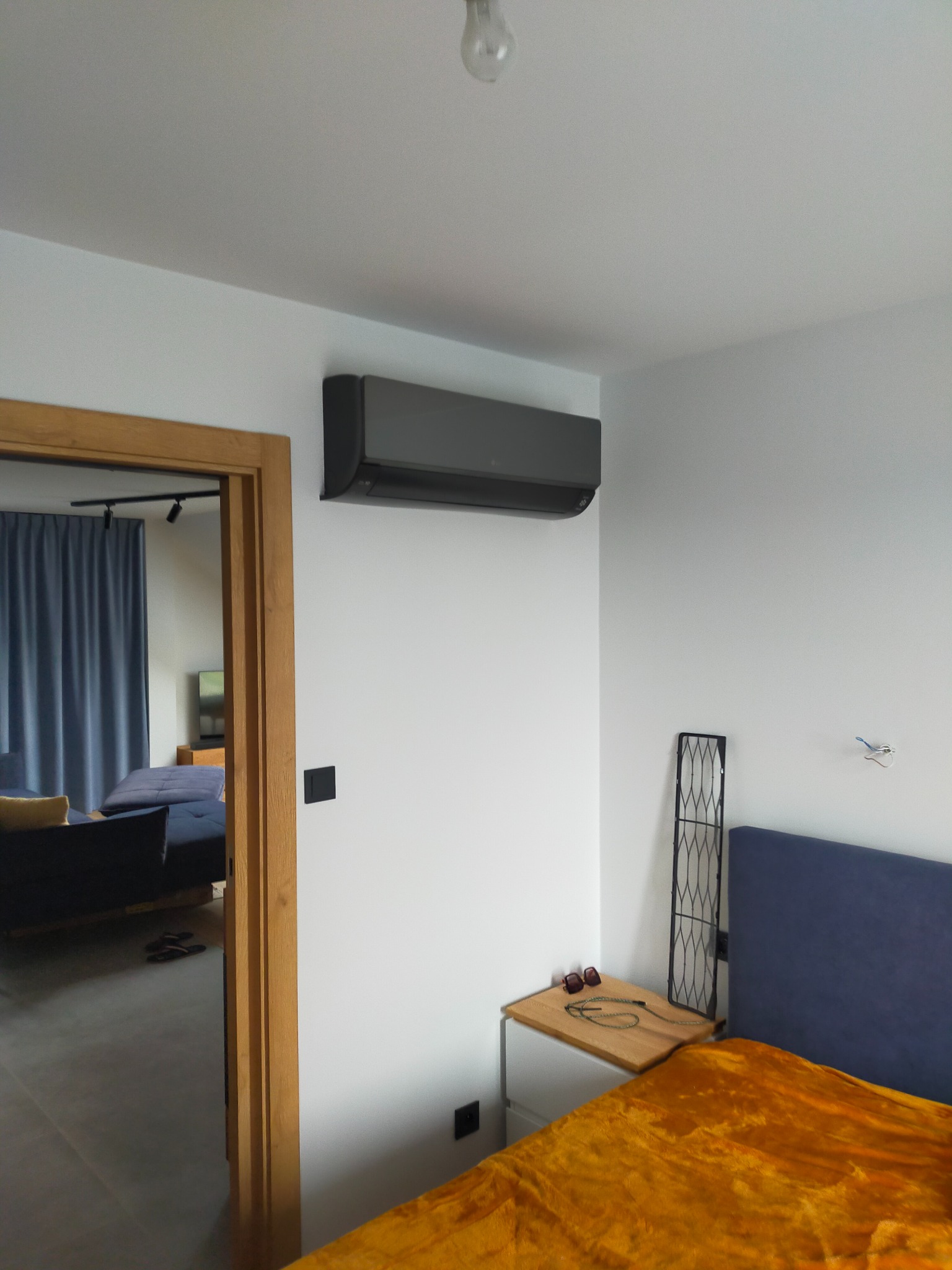 Szary klimatyzator ścienny w sypialni z żółtą narzutą, widok przez otwarte drzwi na pokój z niebieskimi zasłonami i kanapą. Nowoczesny design.