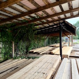 Drewno Chyliczki - Zadaszony skład tarcicy budowlanej z drewnianą konstrukcją, blachą trapezową i zieloną roślinnością. Ułożone deski w równych stosach, widok z perspektywy.