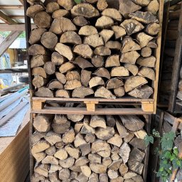 Drewno Chyliczki - Rzędami ułożone drewno kominkowe w drewnianej skrzyni na palecie. Widoczne sęki i pęknięcia drewna. W tle zarys budynku gospodarczego.