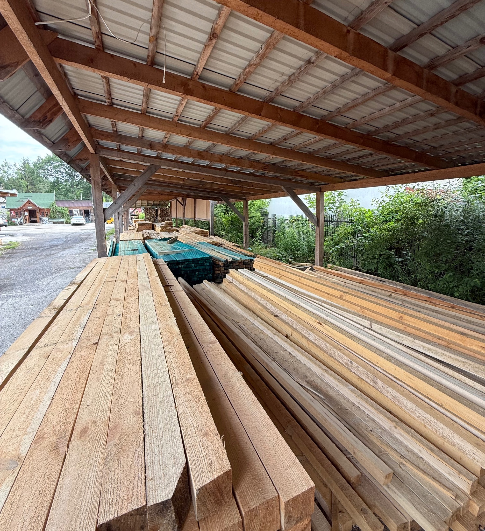 Skład drewna: stosy tarcicy budowlanej pod zadaszeniem, widoczne różne odcienie drewna, w tle zieleń. Drewno ułożone równolegle, tworząc uporządkowany widok.