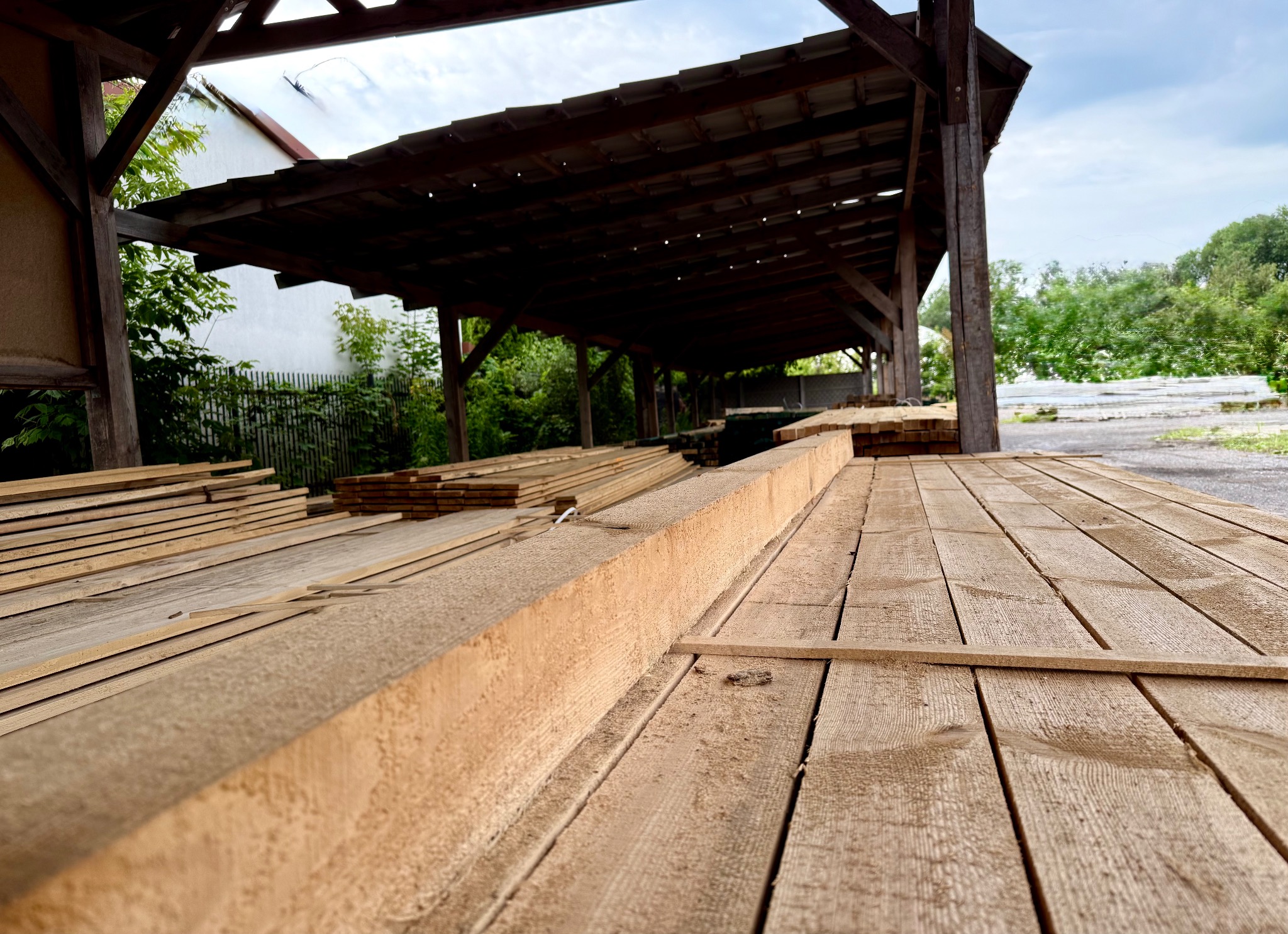 Ujęcie z dołu na składowane deski tarcicy budowlanej pod zadaszeniem, widoczne słoje drewna i elementy konstrukcji dachu. W tle zieleń drzew i fragment budynku.