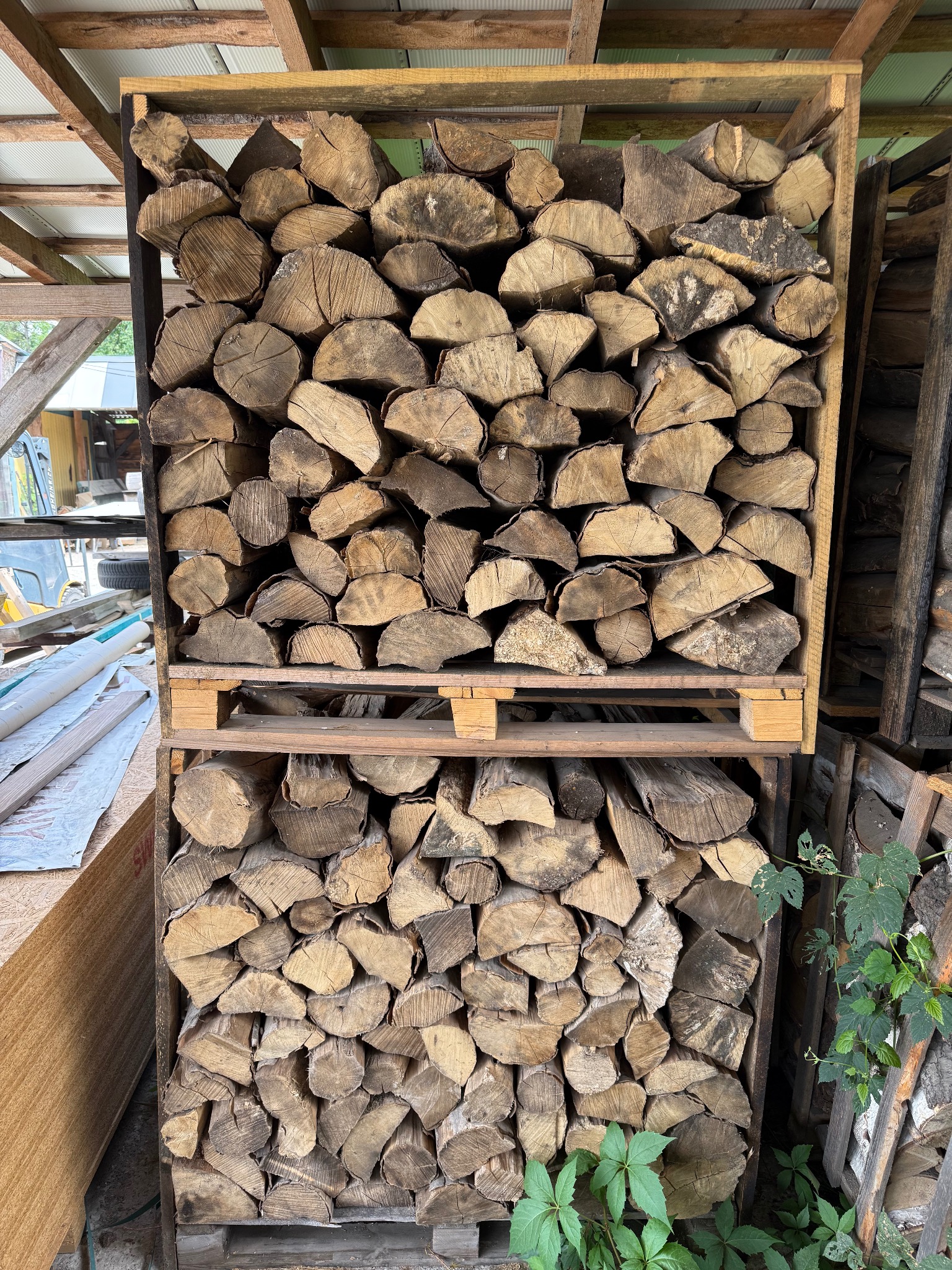 Rzędami ułożone drewno kominkowe w drewnianej skrzyni na palecie. Widoczne sęki i pęknięcia drewna. W tle zarys budynku gospodarczego.