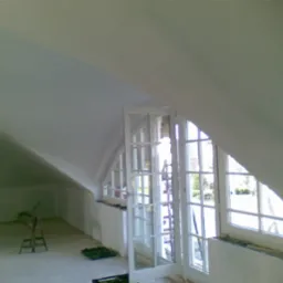 Poddasze ze skośnym sufitem i oknami, ściany po szpachlowaniu, przygotowane do malowania, widoczne narzędzia malarskie i drabina.