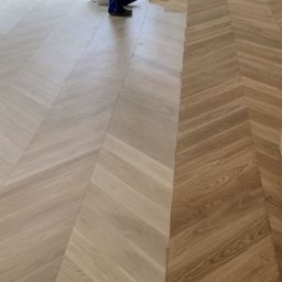 MS Skrzypczak Sp. z o.o. - Parkiet ułożony we wzór jodełki, po prawej stronie w odcieniu naturalnego drewna, po lewej jaśniejszy, zabezpieczony taśmą malarską. Widoczne fragmenty osoby.