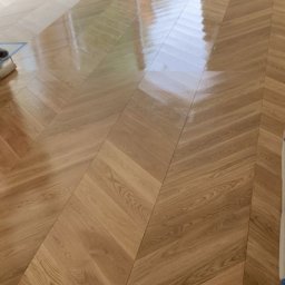 MS Skrzypczak Sp. z o.o. - Lakierowanie parkietu ułożonego w jodełkę. Widoczny wałek i taśma malarska zabezpieczająca listwy przypodłogowe. Część podłogi jest już polakierowana.