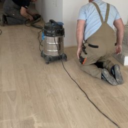 MS Skrzypczak Sp. z o.o. - Prace wykończeniowe: polerowanie jasnej, drewnianej podłogi w pomieszczeniu z surowymi schodami i odkurzaczem przemysłowym, wykonywane przez dwóch fachowców.