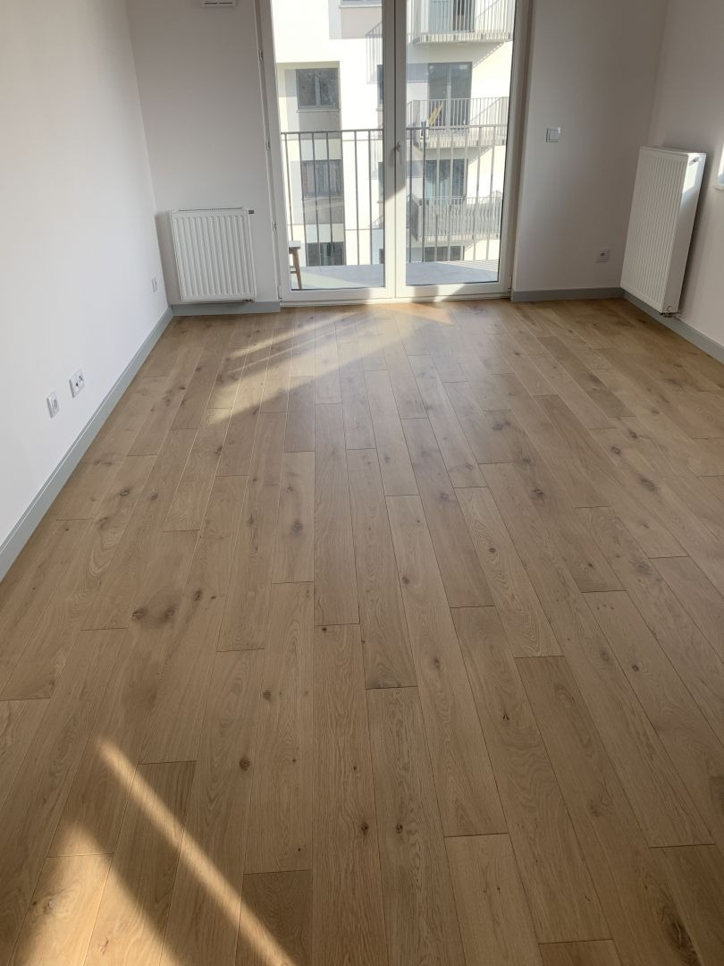 Jasny, drewniany parkiet w nowym mieszkaniu z balkonem. Świeżo położona podłoga, widoczne promienie słoneczne. W tle grzejnik i okno balkonowe.
