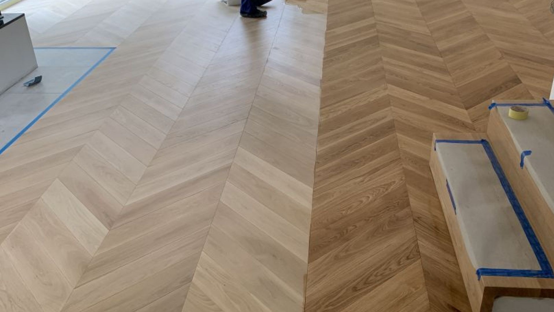 Parkiet ułożony we wzór jodełki, po prawej stronie w odcieniu naturalnego drewna, po lewej jaśniejszy, zabezpieczony taśmą malarską. Widoczne fragmenty osoby.