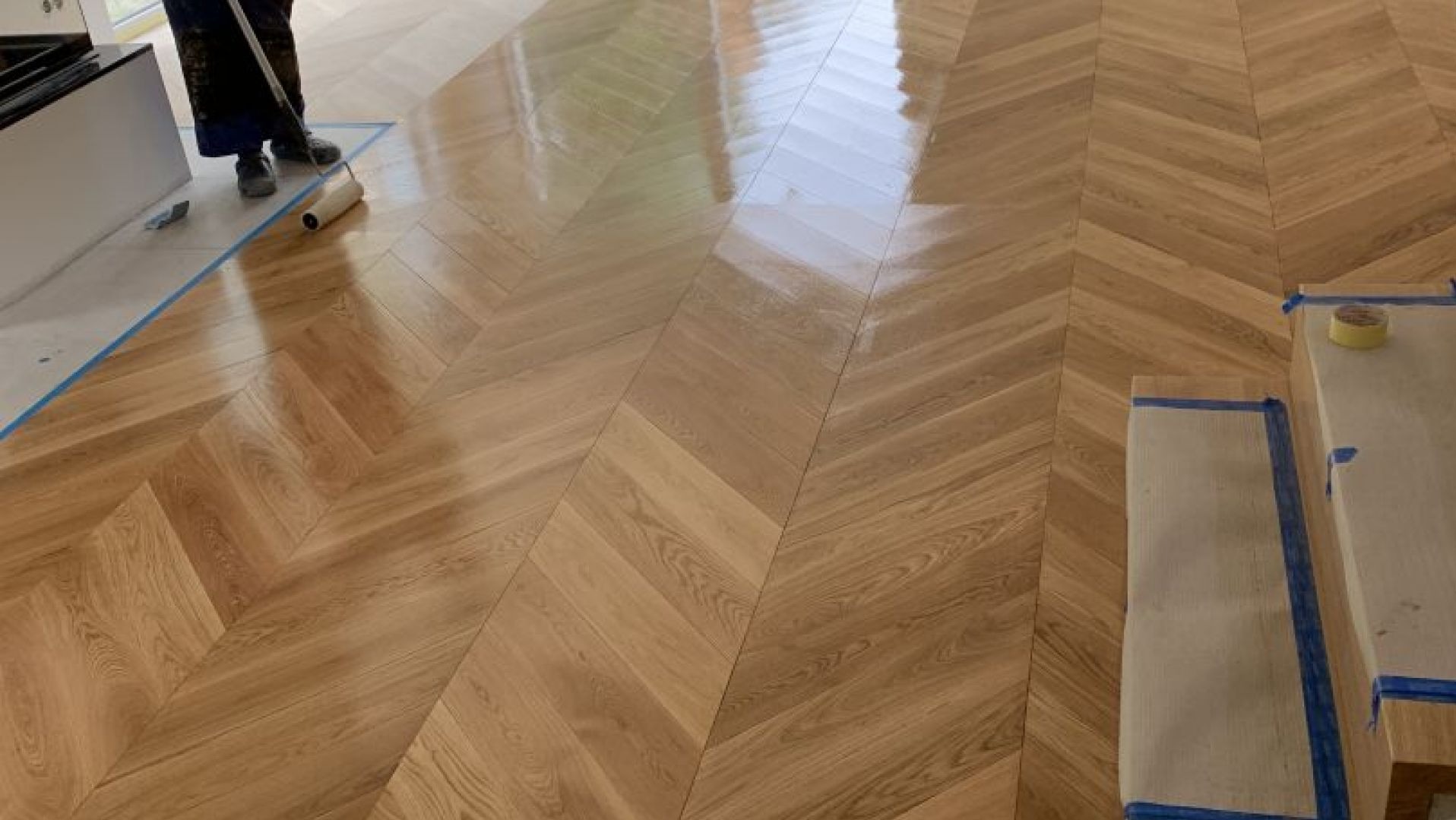 Lakierowanie parkietu ułożonego w jodełkę. Widoczny wałek i taśma malarska zabezpieczająca listwy przypodłogowe. Część podłogi jest już polakierowana.