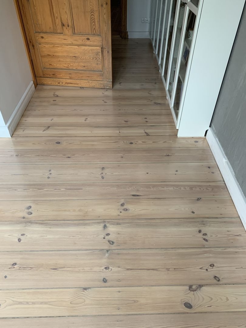 Jasna, odnowiona podłoga z desek w korytarzu, widoczna drewniana stolarka drzwiowa i białe szafy w tle. Naturalne usłojenie drewna.