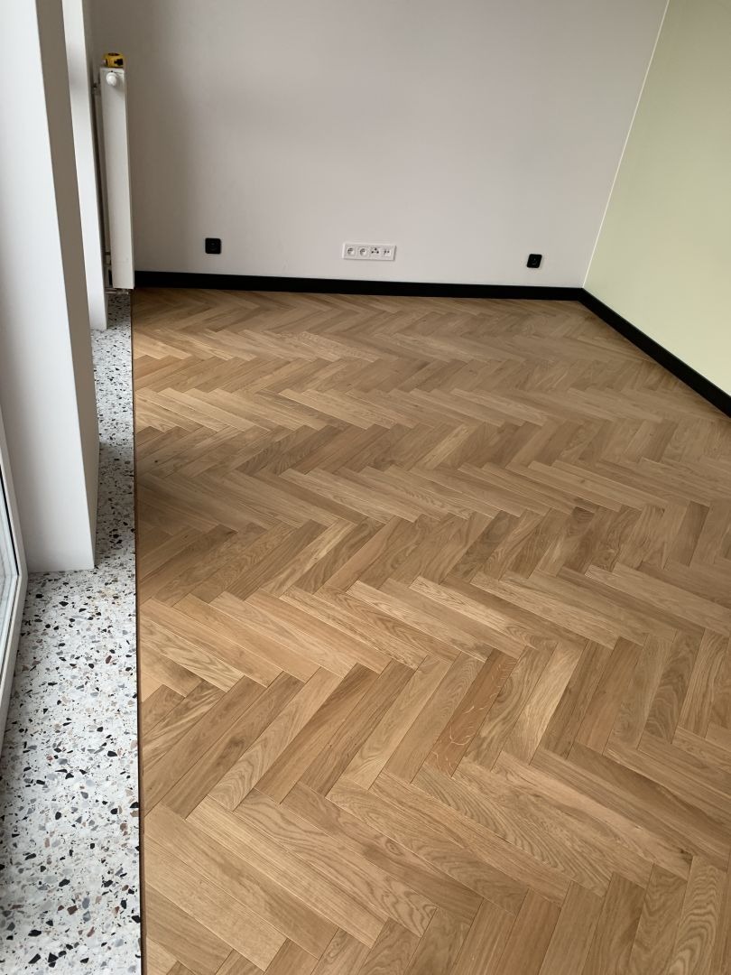 Parkiet ułożony we wzór jodełki francuskiej, z widocznymi słojami drewna, kontrastujący z lastryko przy oknie i czarną listwą przypodłogową.