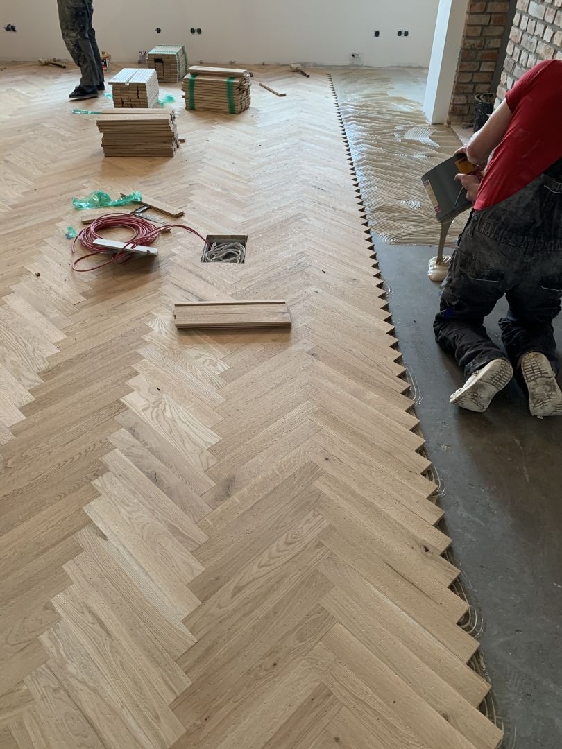 Układanie parkietu w jodełkę: widok z góry na klejony parkiet, częściowo ułożony, z przygotowanym klejem i narzędziami. W tle paczki z deskami.