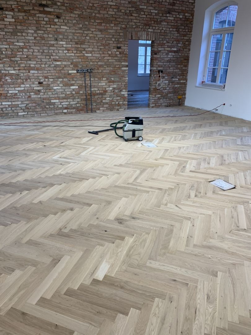 Jasny parkiet ułożony w jodełkę francuską w przestronnym wnętrzu z surową cegłą. Widoczny odkurzacz przemysłowy i notatnik. Duże okno w tle.