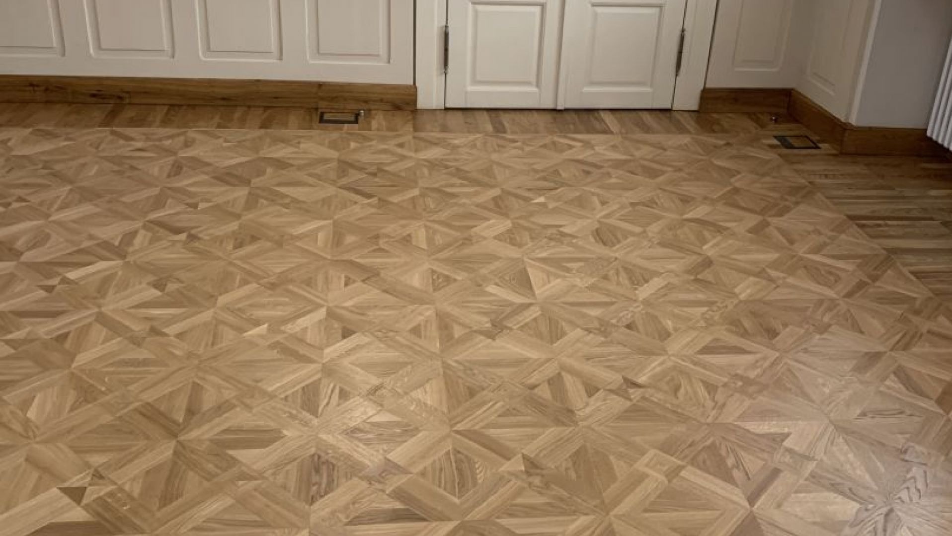Parkiet ułożony we wzór geometryczny, jasne drewno, widok z góry na podłogę w pokoju z białymi drzwiami i listwami przypodłogowymi.