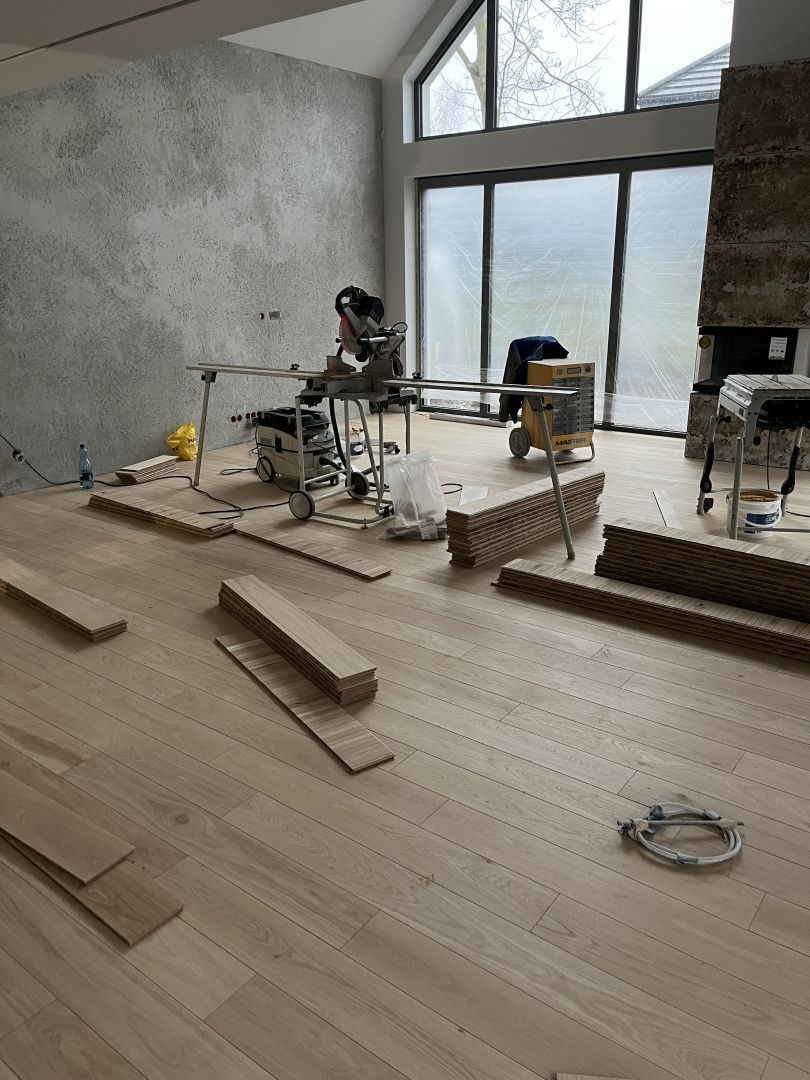 Jasna drewniana podłoga w trakcie montażu. Widoczne deski, narzędzia i maszyny do obróbki drewna w przestronnym, jasnym pomieszczeniu z dużymi oknami i surowym wykończeniem ścian.