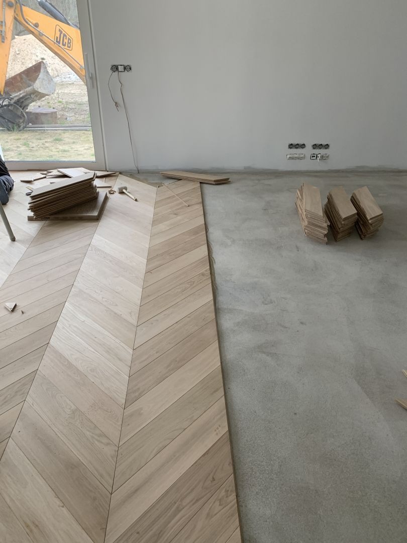 Układanie parkietu w jodełkę francuską, widoczne deski i część surowej posadzki. W tle okno z widocznym sprzętem budowlanym.