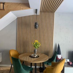 ProMal - Nowoczesny salon z drewnianymi lamelami na ścianie i suficie, okrągły stół z żółtymi tulipanami, welurowe krzesła w odcieniach zieleni i złota. Minimalistyczny design.