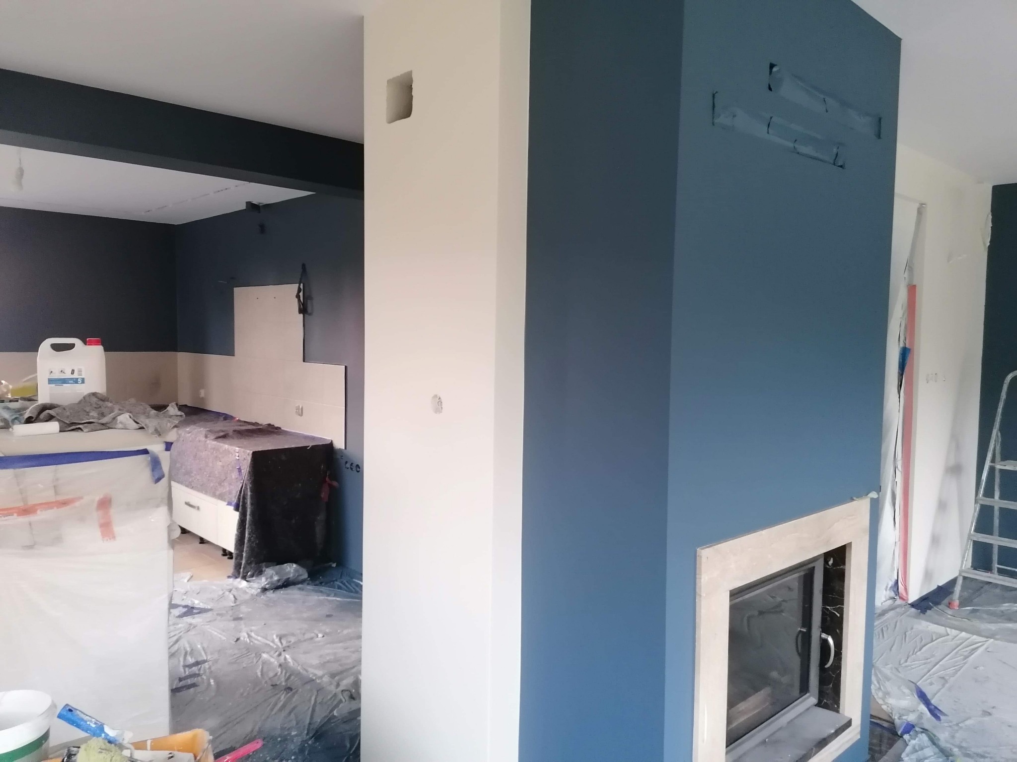 Świeżo pomalowane wnętrze z fragmentem kominka, zabezpieczone folią meble i podłoga. Widoczne ślady po taśmie malarskiej na ścianie.