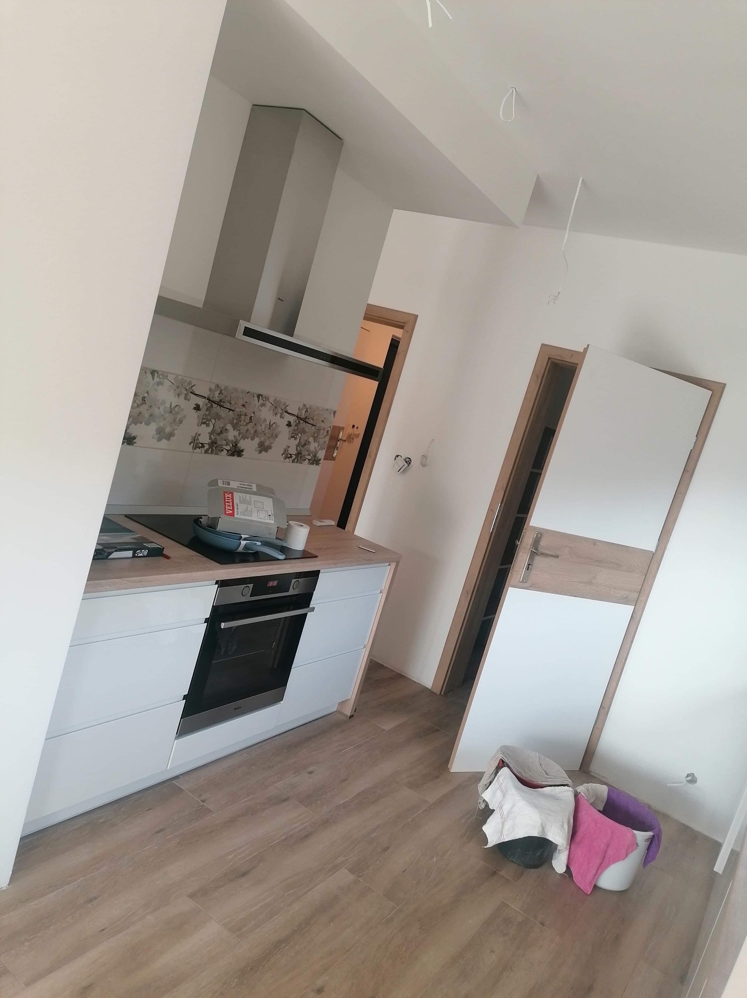 Kuchnia po remoncie: białe szafki, drewnopodobne panele, okap i płytki z motywem kwiatowym. Widoczne ślady po remoncie, np. wiadro z szmatami.