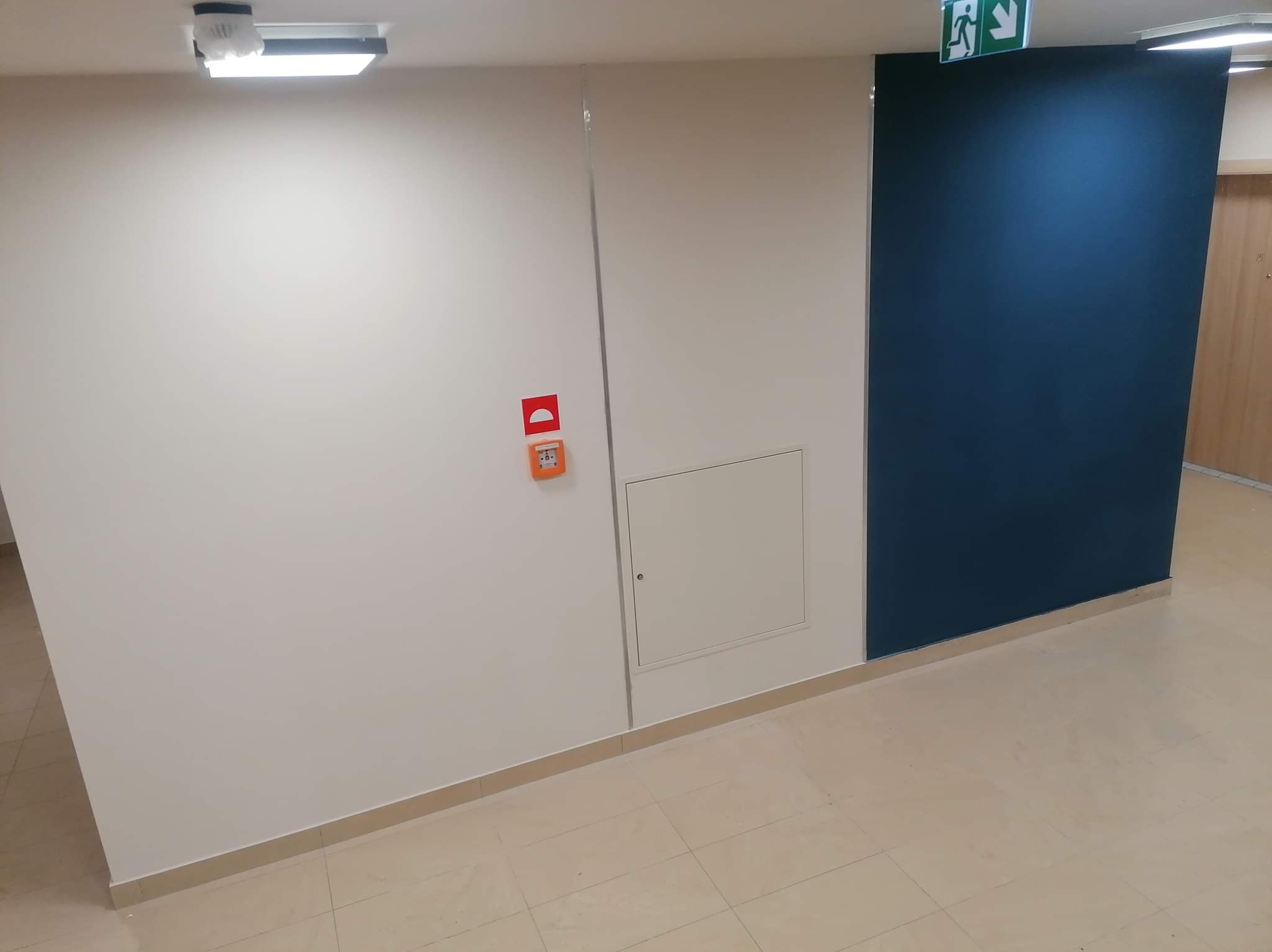 Korytarz po malowaniu: białe ściany z alarmem przeciwpożarowym, granatowa ściana z piktogramem ewakuacyjnym, beżowa podłoga z płytek. Widok z dołu.