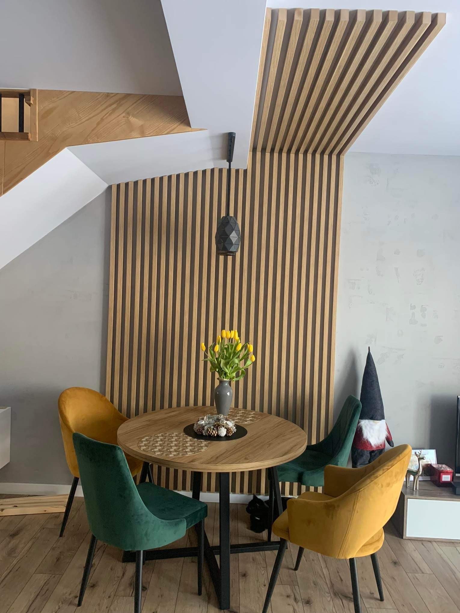 Nowoczesny salon z drewnianymi lamelami na ścianie i suficie, okrągły stół z żółtymi tulipanami, welurowe krzesła w odcieniach zieleni i złota. Minimalistyczny design.