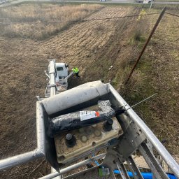 ELEKTROINVEST Sp. z o.o. - Widok z podnośnika koszowego na kabel w opakowaniu, narzędzia i pracownika w odblaskowej kamizelce, przygotowującego się do pracy na wysokości.