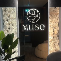 ELEKTROINVEST Sp. z o.o. - Nowoczesne oświetlenie LED wkomponowane w kamienną ścianę, widoczne przez szklane drzwi z logo 'Muse', z elementami roślinnymi i meblami w tle.