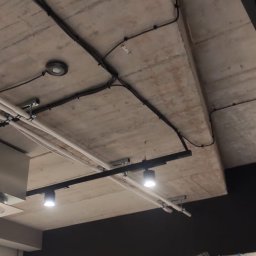 ELEKTROINVEST Sp. z o.o. - Betonowy sufit z widocznymi czarnymi kablami elektrycznymi, punktowym oświetleniem i szynami. Częściowo widoczne drzwi zabezpieczone folią.