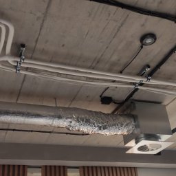 ELEKTROINVEST Sp. z o.o. - Betonowy sufit z widocznymi instalacjami elektrycznymi: czarne i białe kable, oświetlenie punktowe oraz fragment instalacji wentylacyjnej. Widok z dołu.