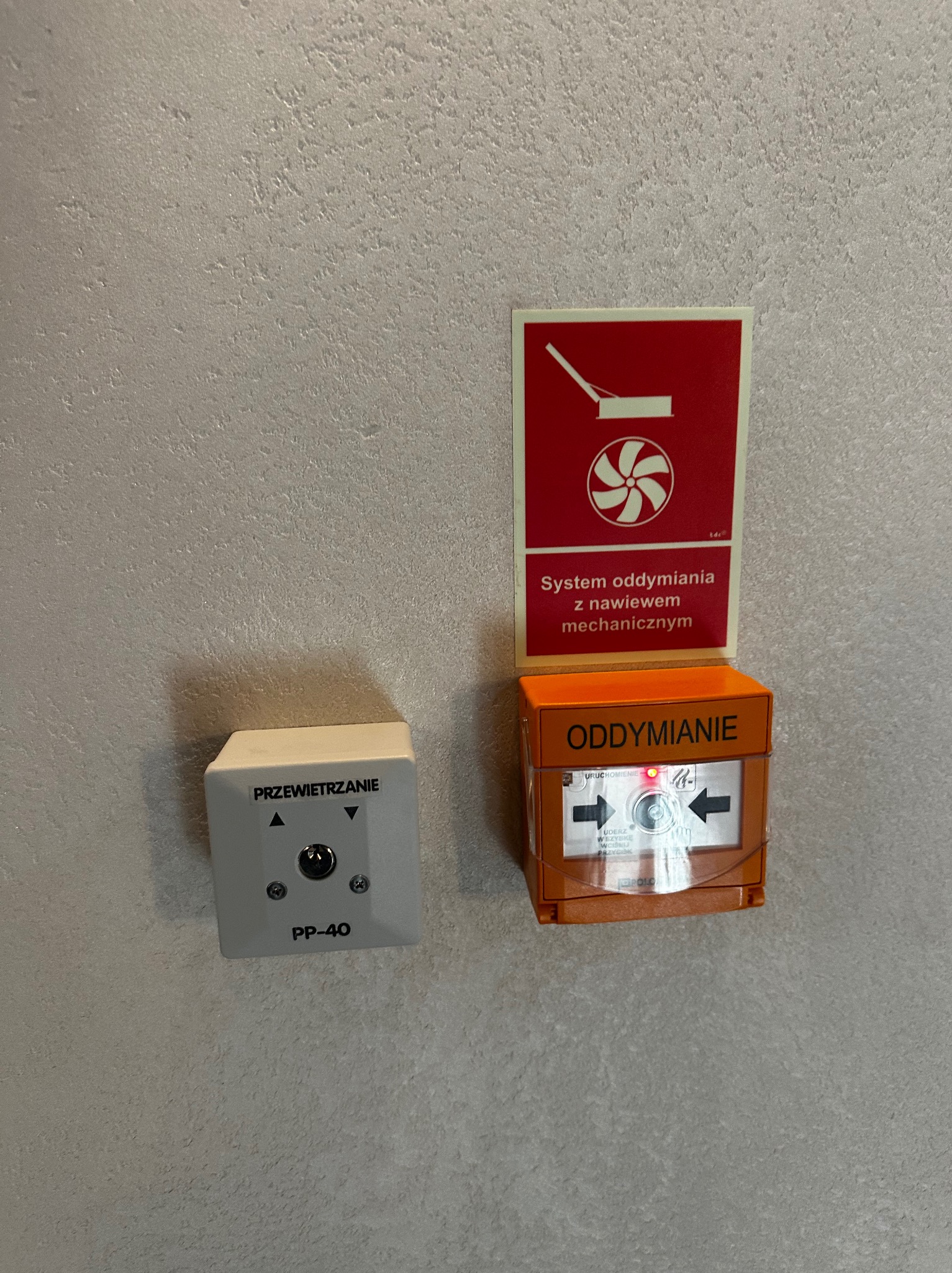 Przycisk oddymiania w pomarańczowej obudowie, obok przycisk wentylacji PP-40 i czerwona tabliczka informacyjna systemu oddymiania z nawiewem mechanicznym na szarej ścianie.