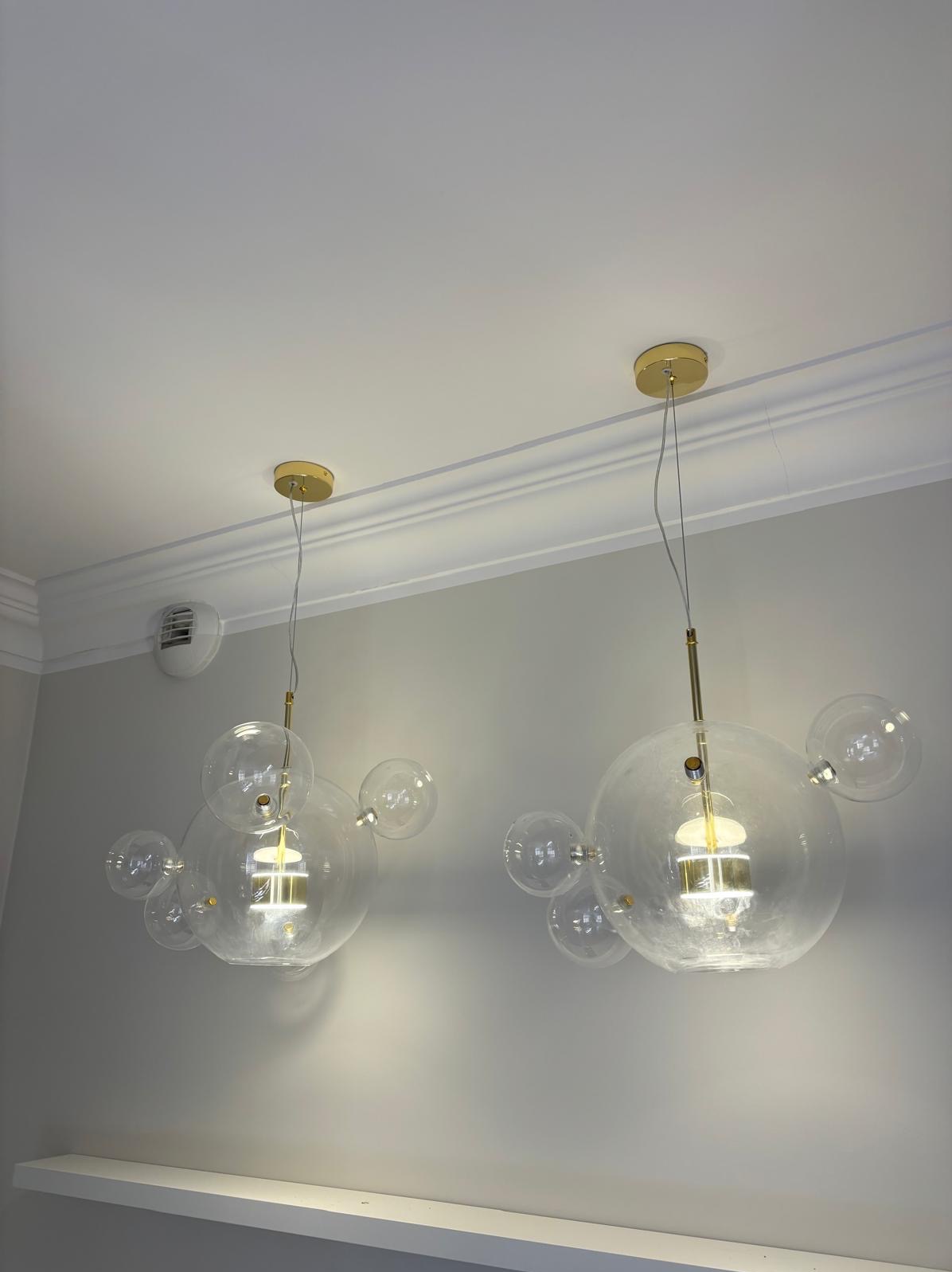 Dwa nowoczesne, szklane żyrandole w kształcie kuli z mniejszymi kulkami, zawieszone na złotych podsufitkach, oświetlające wnętrze. Minimalistyczny design.