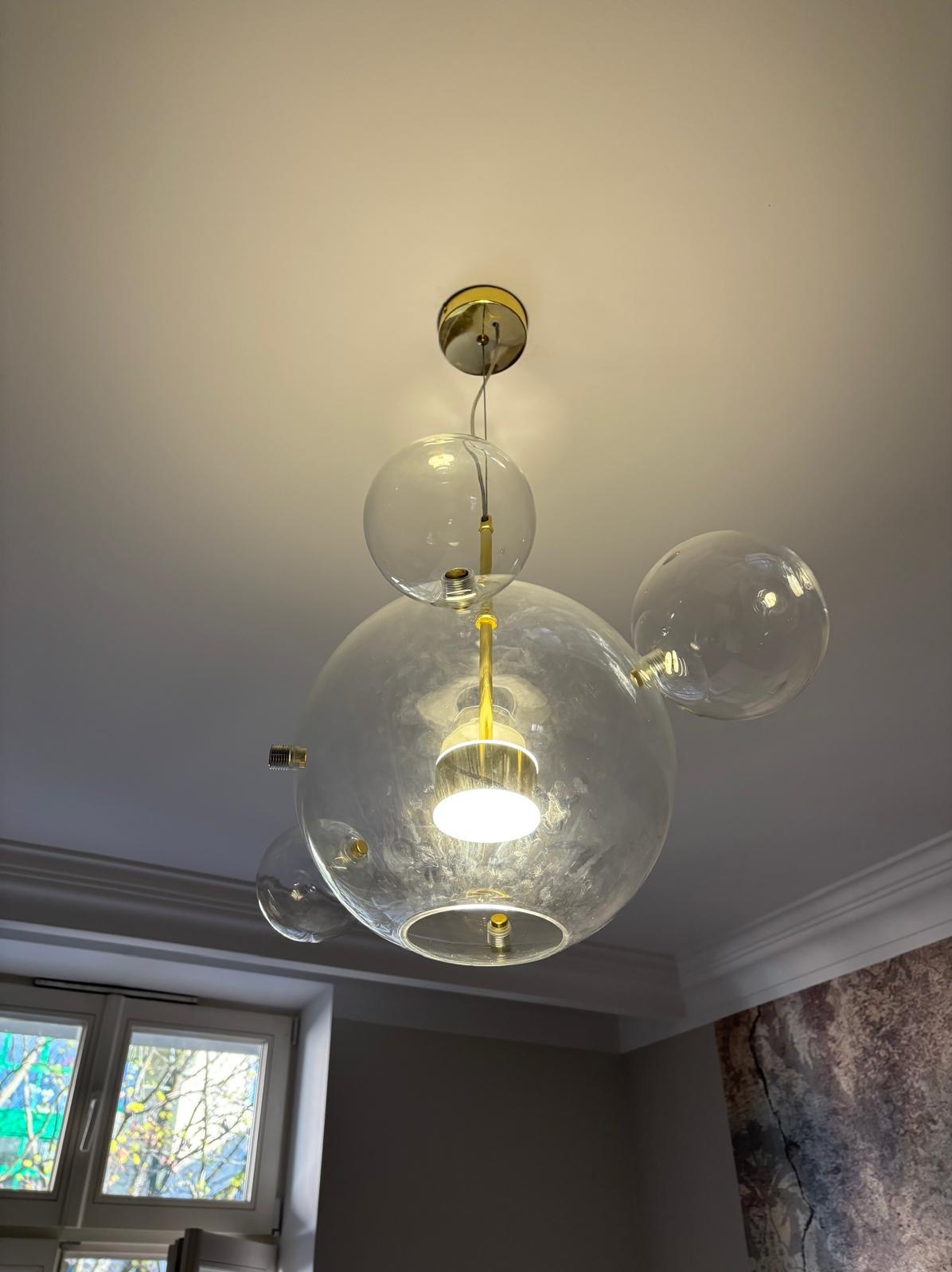 Szklana lampa sufitowa w kształcie kuli z złotymi elementami, oświetlająca wnętrze. Widok od dołu, okno w tle.