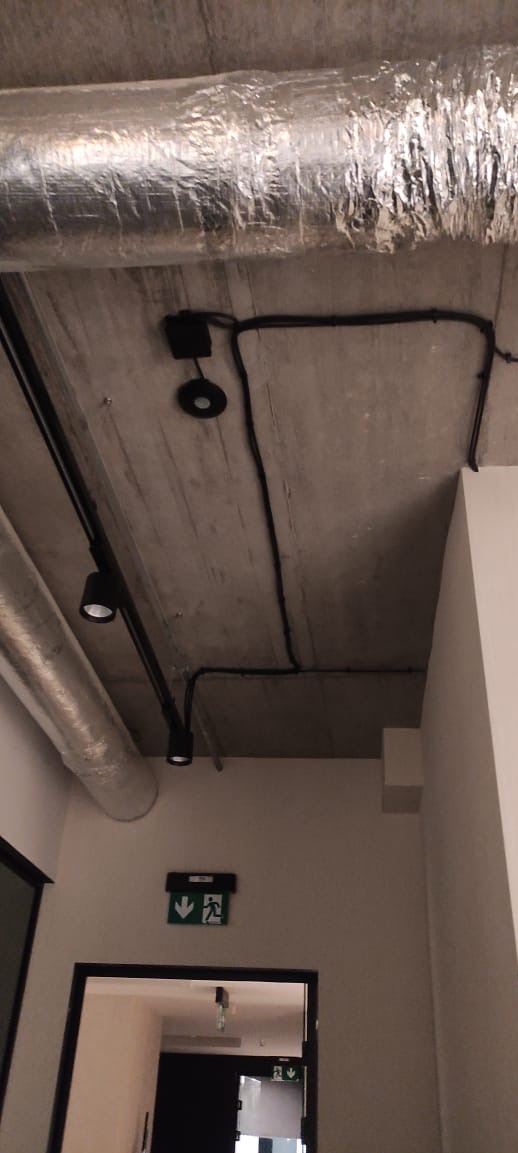 Betonowy sufit z widocznymi przewodami elektrycznymi, czarnymi reflektorami na szynie i rurami wentylacyjnymi. Widoczny znak ewakuacyjny nad drzwiami.