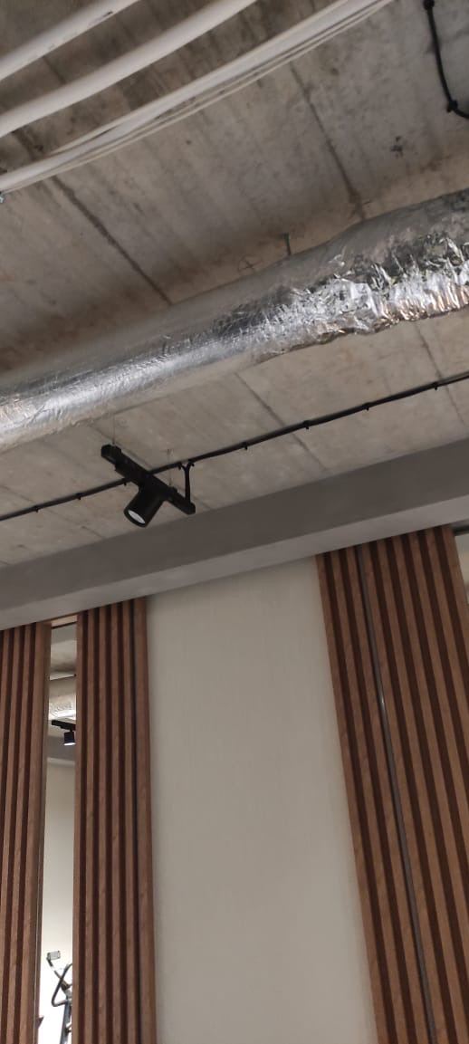 Szyna z reflektorem punktowym na suficie betonowym, widoczne rury wentylacyjne w folii aluminiowej, drewniane lamele dekoracyjne na ścianie.