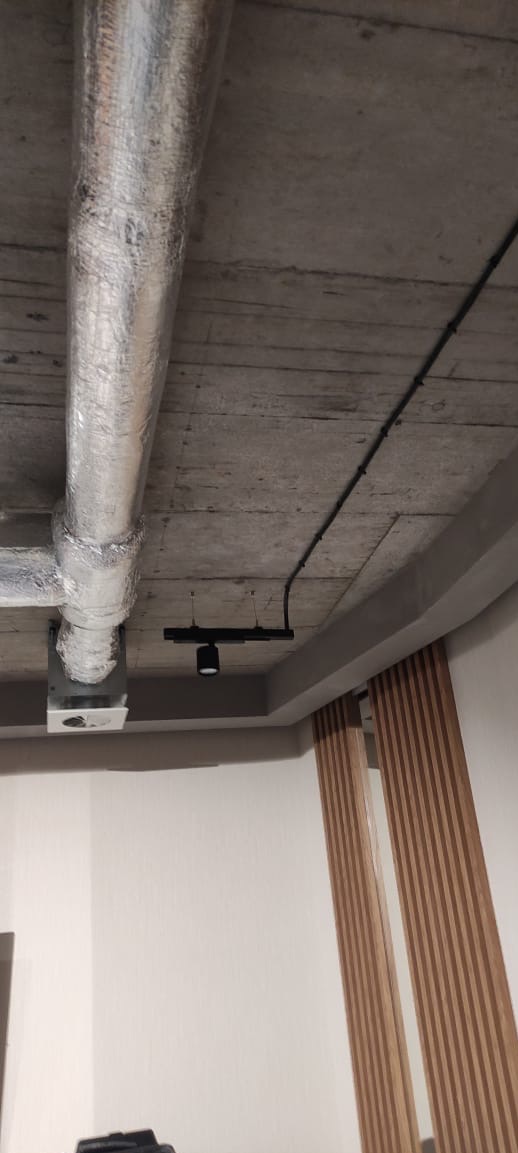 Betonowy sufit z widoczną instalacją wentylacyjną w srebrnej izolacji, czarną listwą oświetleniową i przewodami elektrycznymi. Elementy dekoracyjne z drewna na ścianie.