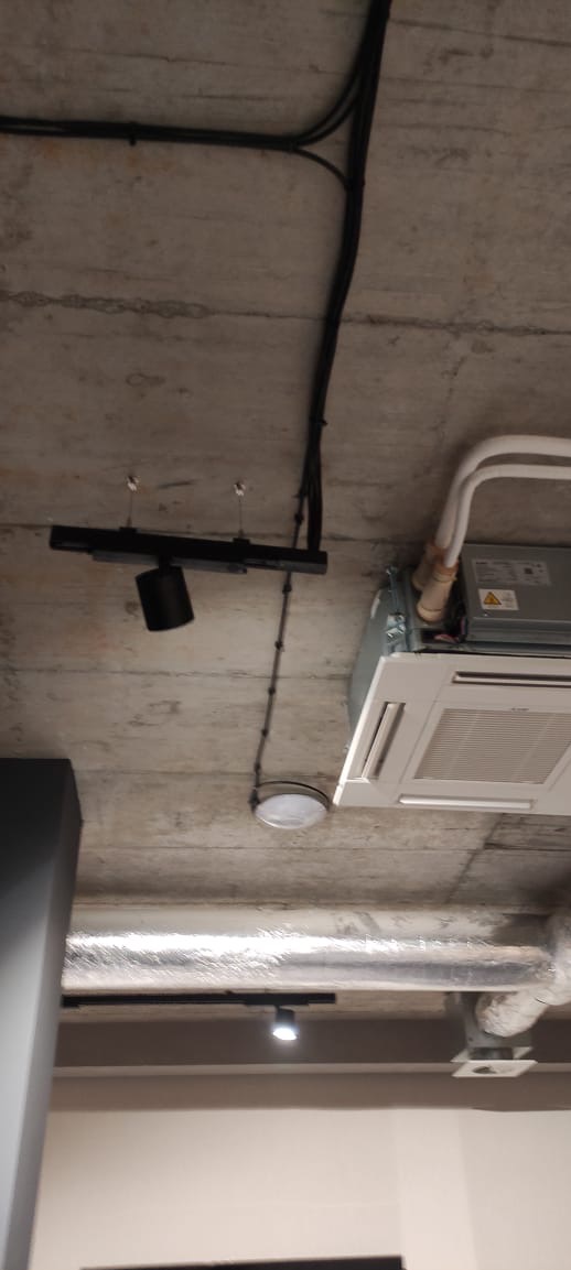 Betonowy sufit z widocznymi instalacjami: czarne kable, szyna z reflektorem punktowym, okrągła lampa sufitowa i klimatyzator. Surowy, industrialny styl.