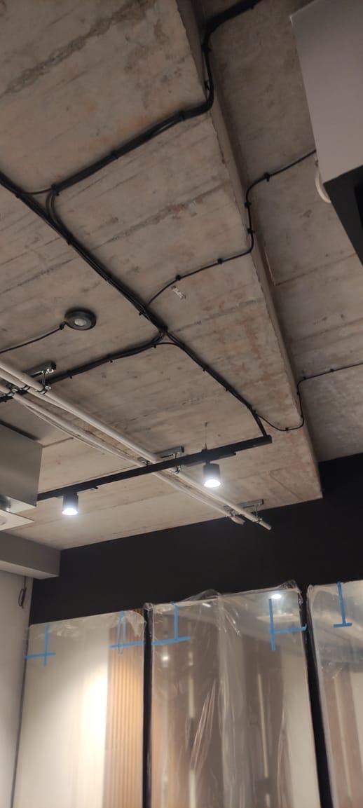 Betonowy sufit z widocznymi czarnymi kablami elektrycznymi, punktowym oświetleniem i szynami. Częściowo widoczne drzwi zabezpieczone folią.