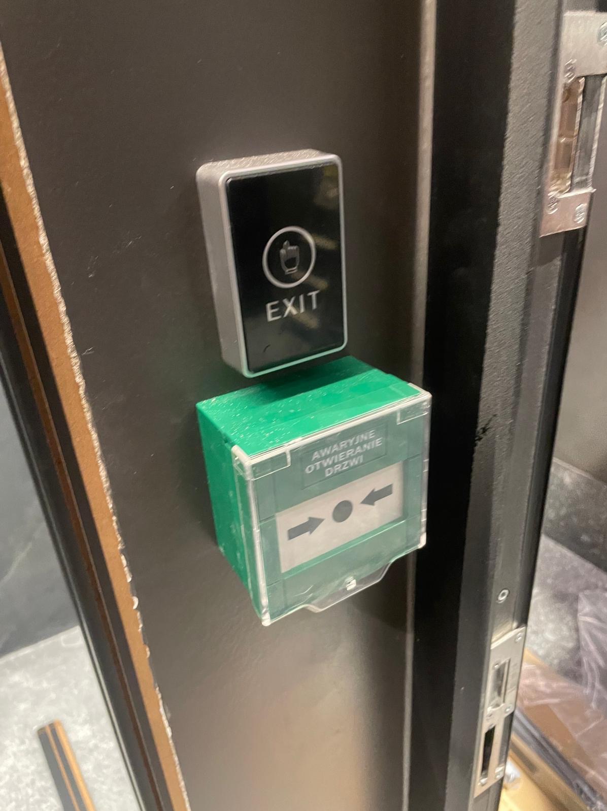 Przycisk wyjścia awaryjnego z zieloną obudową i napisem 'Awaryjne Otwieranie Drzwi' zamontowany na ciemnej ścianie obok framugi drzwi.