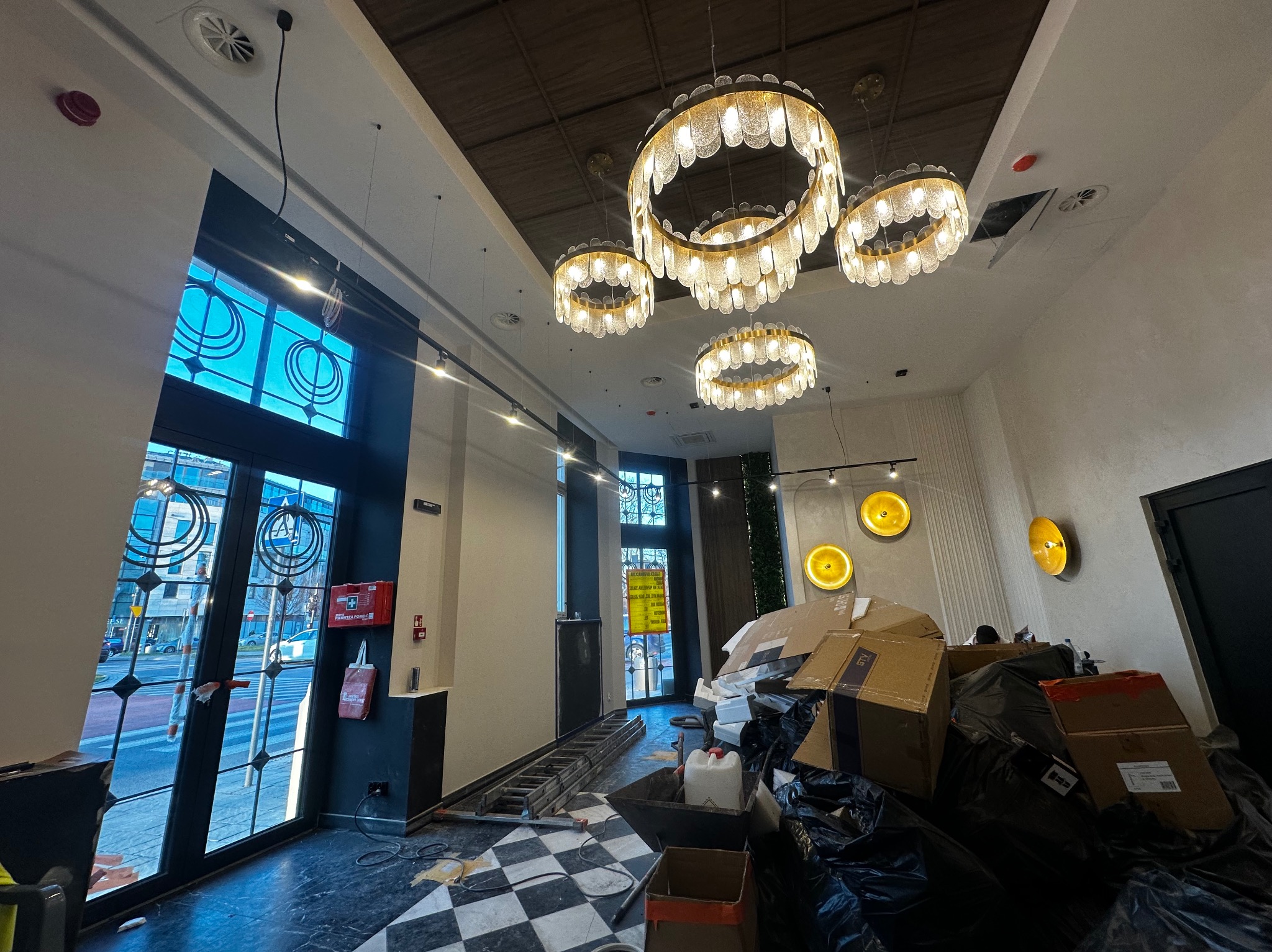 Wnętrze lokalu w trakcie montażu oświetlenia: widoczne wiszące, okrągłe lampy z dekoracyjnymi elementami, drabina, kartony i folie zabezpieczające podłogę.